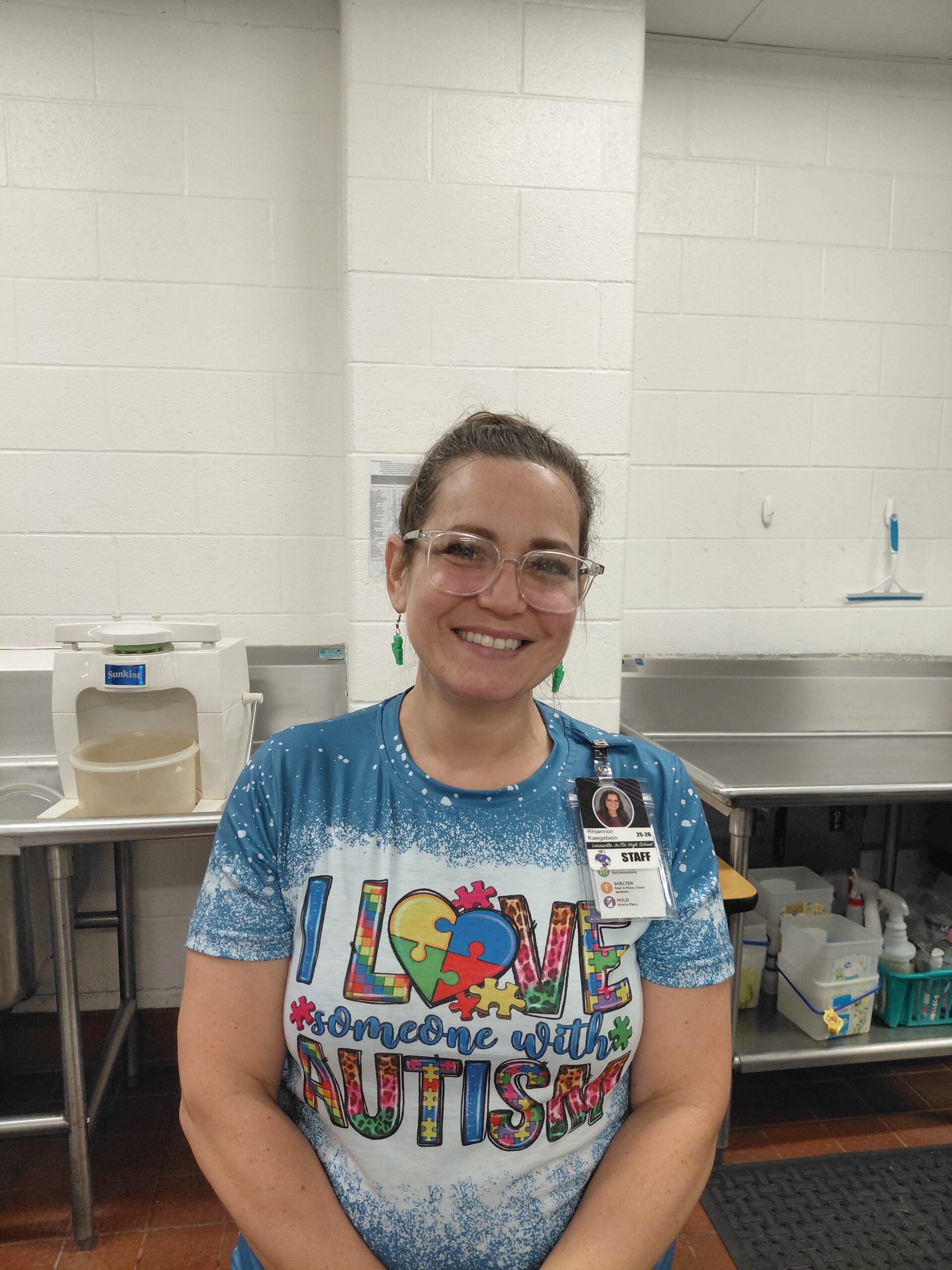 Smiling female staff member with glasses and a name badge in a stainless steel kitchen area behind her.