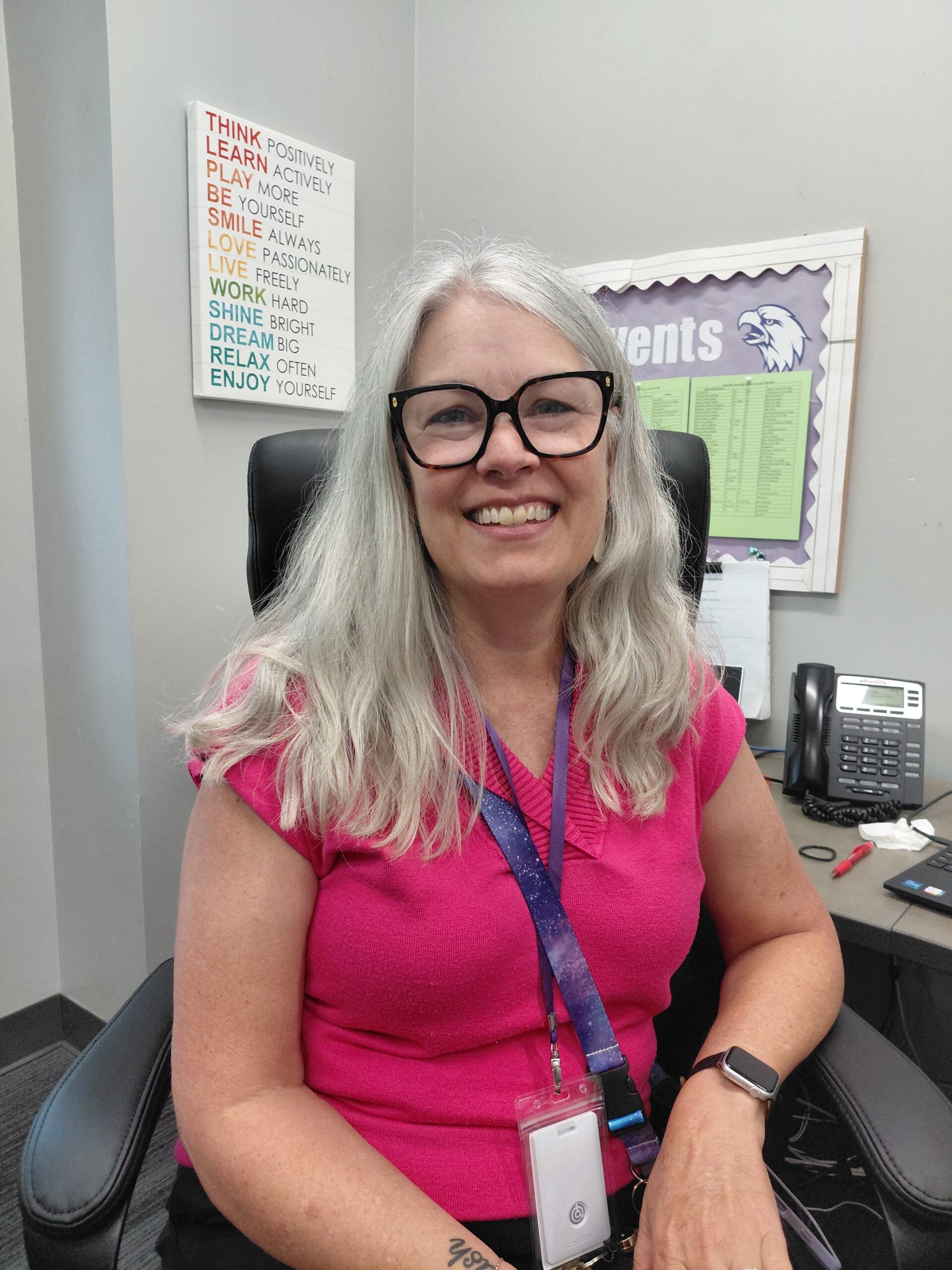 Smiling woman with gray hair and black glasses sits in an office, wearing a pink top and purple lanyard badge.