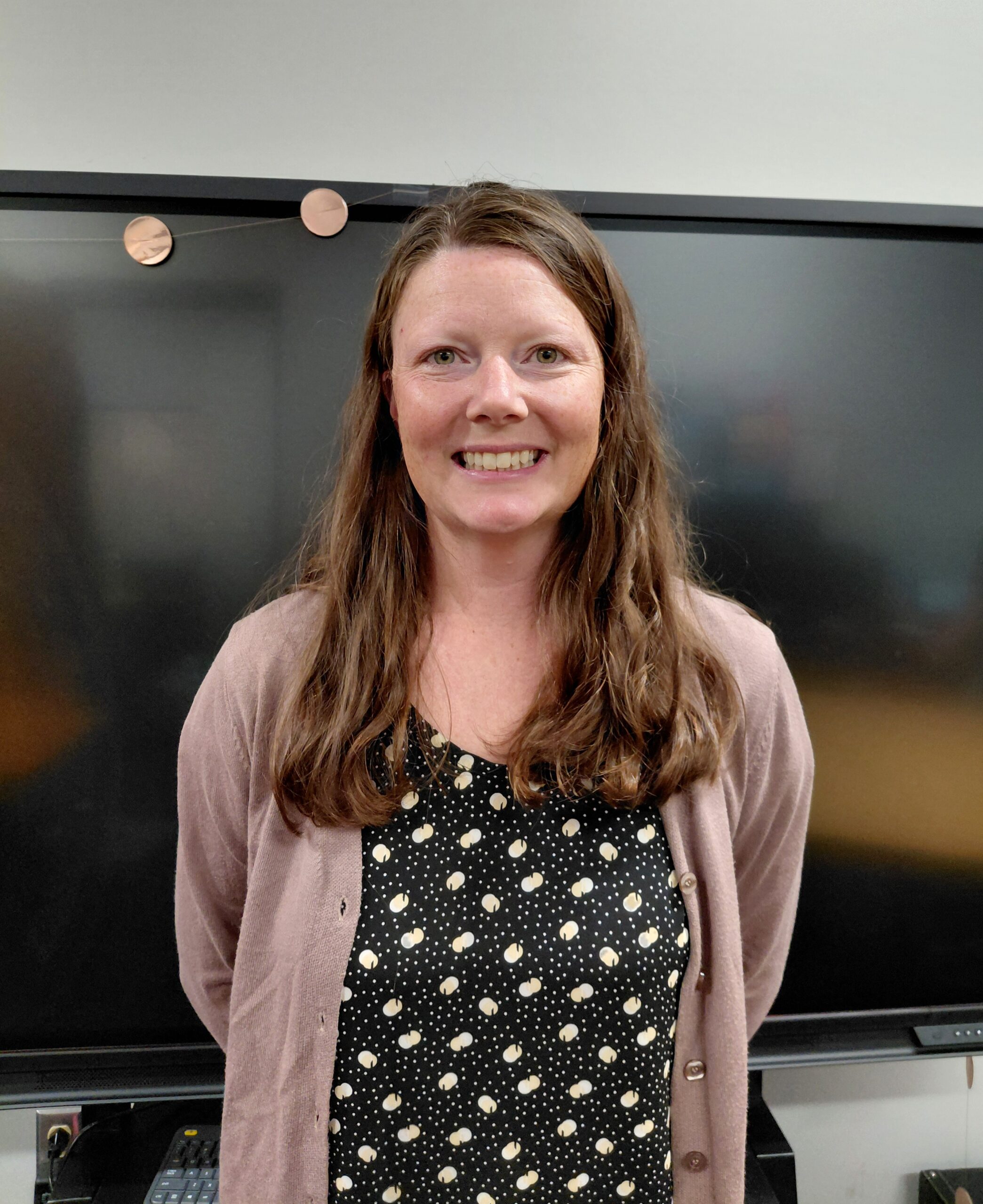 Smiling woman with long brown hair wearing a pink cardigan over a black polka-dot dress, standing in front of a dark screen.
