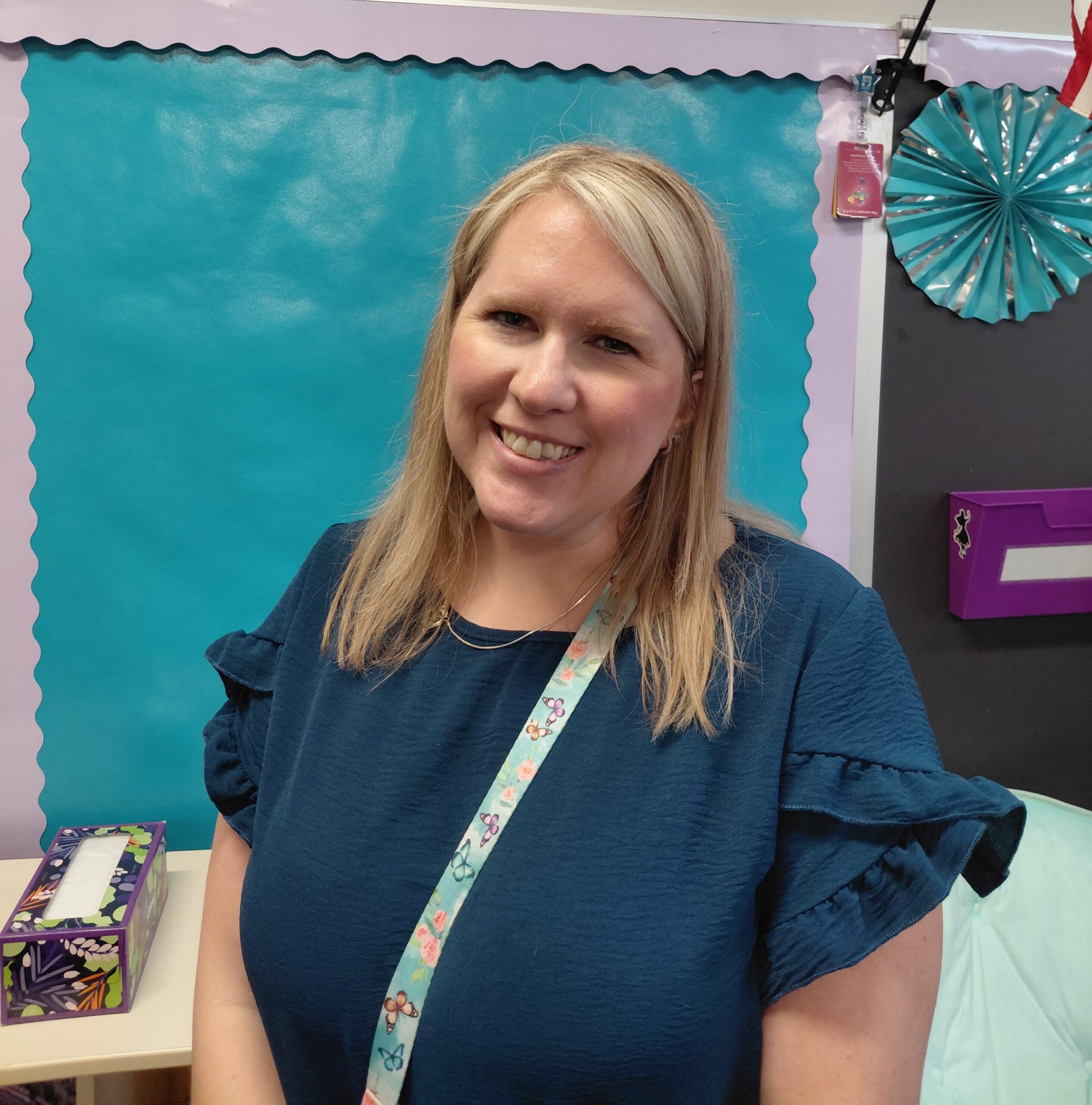 Close-up selfie of a smiling woman wearing a blue top, with a butterfly-pattern lanyard in a classroom setting.